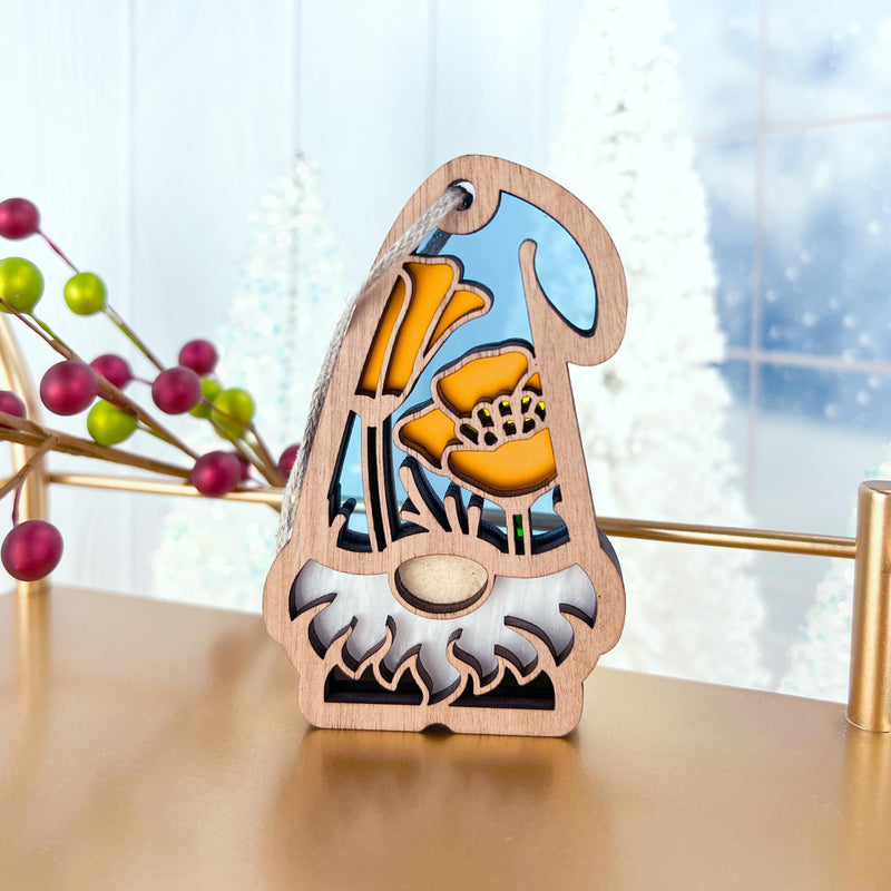 The California Poppy Ornament by Forged Flare®—a 3.75" Nature's Gnomes™ garden gnome with a white beard, yellow flowers, and sky-blue background—stands on a table amid blurred frosted trees and decorative berries.