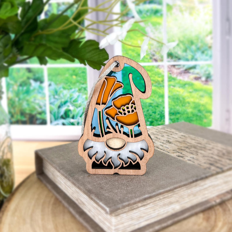 The Forged Flare® California Poppy Ornament, a 3.75" garden gnome from Nature's Gnomes™, features a white beard and orange flower hat, perfect for displaying on stacked books by your window with a garden view.
