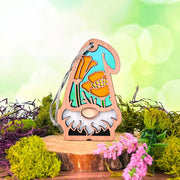 The Forged Flare® California Poppy Ornament is a 3.75" tall garden gnome from the Nature's Gnomes™ collection, featuring an orange flower-painted hat, standing on a wood slice with moss and pink blooms, set against a green bokeh background.
