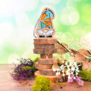 The 3.75" California Poppy Ornament garden gnome from Forged Flare’s Nature’s Gnomes™ stands on stacked wood discs, surrounded by colorful flowers, dried leaves, white berries, and purple-green moss against a blurred green-blue backdrop.