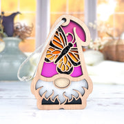 The Forged Flare® Monarch Butterfly Ornament, a 3.75" wooden garden gnome from the Nature's Gnomes™ collection, features a pink hat with a butterfly design and sits on a white surface with blurred vases and flowers behind it.