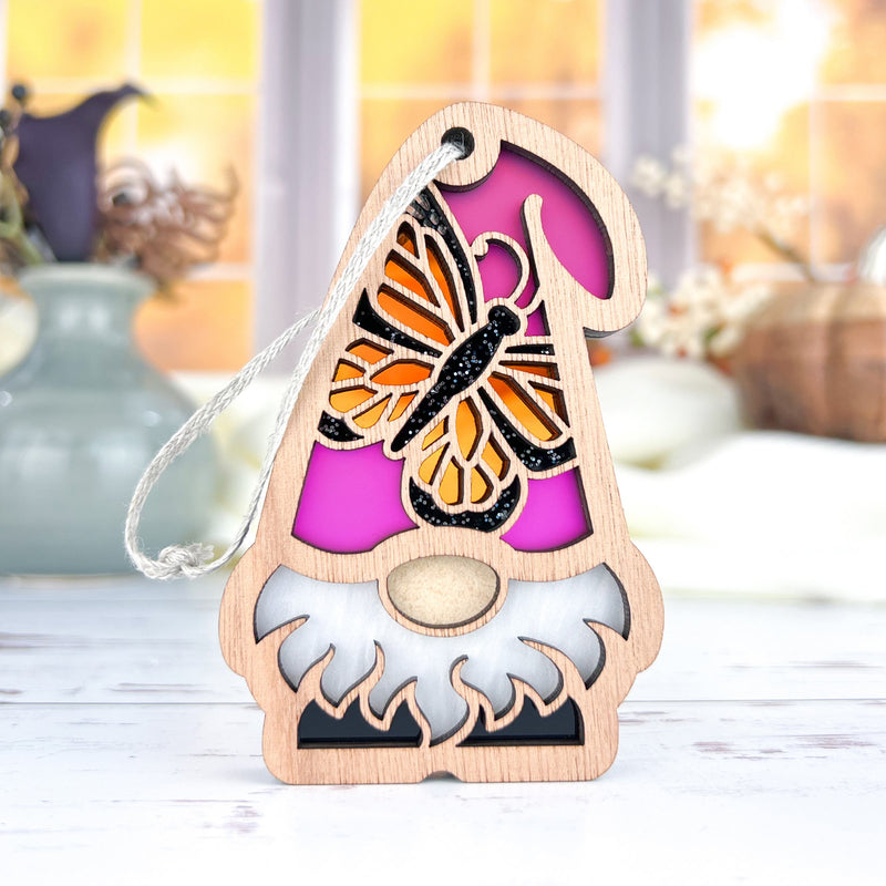 The Forged Flare® Monarch Butterfly Ornament, a 3.75" wooden garden gnome from the Nature's Gnomes™ collection, features a pink hat with a butterfly design and sits on a white surface with blurred vases and flowers behind it.
