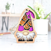 The Forged Flare® Monarch Butterfly Ornament is a 3.75" wooden garden gnome with a white beard, pink hat, and butterfly cutout from the Nature’s Gnomes™ collection, displayed in a bright room with a potted plant and blurred decor.