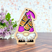 The Forged Flare® Monarch Butterfly Ornament, a 3.75" garden gnome from the Nature’s Gnomes™ collection, features a wooden design with a purple hat and butterfly motif, set against purple and white floral accents.