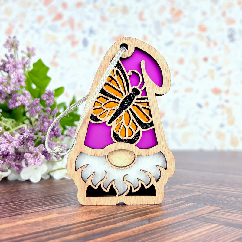 The Forged Flare® Monarch Butterfly Ornament, a 3.75" garden gnome from the Nature’s Gnomes™ collection, features a wooden design with a purple hat and butterfly motif, set against purple and white floral accents.