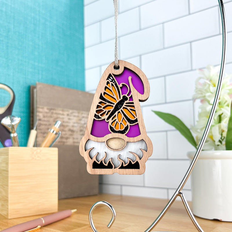 The Forged Flare® Monarch Butterfly Ornament | 3.75" (H) Garden Gnome Gift from Nature's Gnomes™ features a butterfly-adorned hat and hangs by a string, set against a tiled wall, desk organizers with pens, and white flowers in a vase.