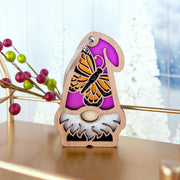 The Monarch Butterfly Ornament by Forged Flare®, a 3.75" wooden gnome from the Nature’s Gnomes™ collection, features a purple hat with an orange monarch butterfly, displayed on gold with red berries and snowy trees in the blurred background.