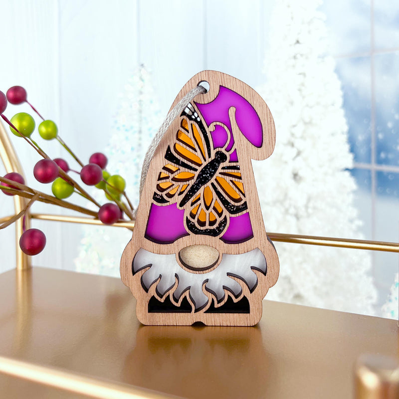 The Monarch Butterfly Ornament by Forged Flare®, a 3.75" wooden gnome from the Nature’s Gnomes™ collection, features a purple hat with an orange monarch butterfly, displayed on gold with red berries and snowy trees in the blurred background.