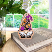 The Forged Flare® Monarch Butterfly Ornament, a 3.75" Nature’s Gnomes™ garden gnome, stands on a closed book with greenery and a sunlit garden visible through the window in the background.
