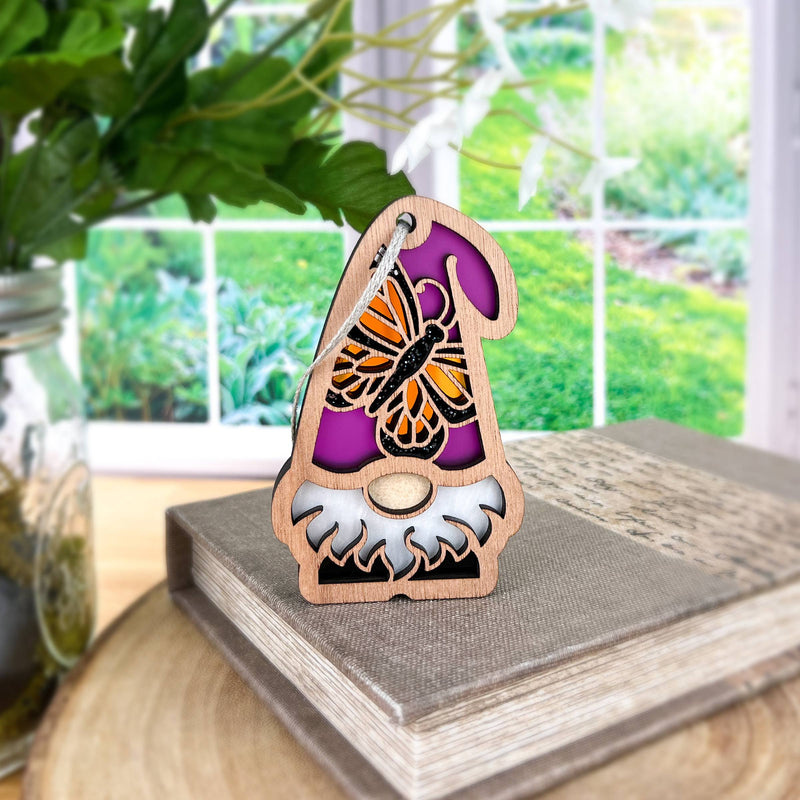 The Forged Flare® Monarch Butterfly Ornament, a 3.75" Nature’s Gnomes™ garden gnome, stands on a closed book with greenery and a sunlit garden visible through the window in the background.