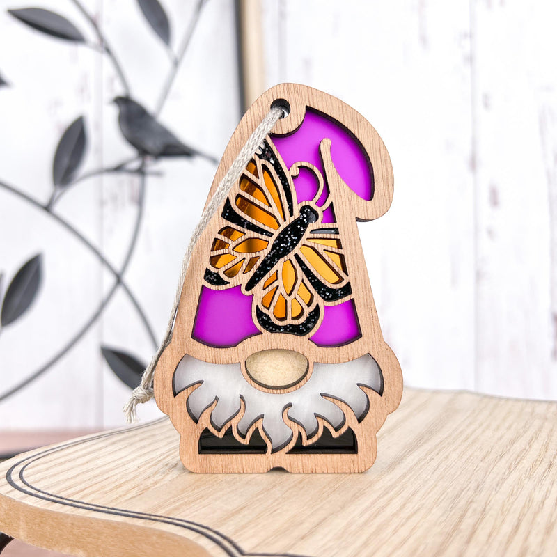 The Forged Flare® Monarch Butterfly Ornament is a 3.75"H wooden gnome with a white beard, pink hat, yellow butterfly, and leafy metal accent. Part of Nature’s Gnomes™, it includes a hanging string—perfect as a garden gnome gift.