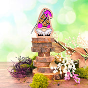 The Forged Flare® Monarch Butterfly Ornament, a 3.75" garden gnome with a pink hat from the Nature’s Gnomes™ collection, stands on stacked wood slices among dried flowers and greenery against a bright green background.