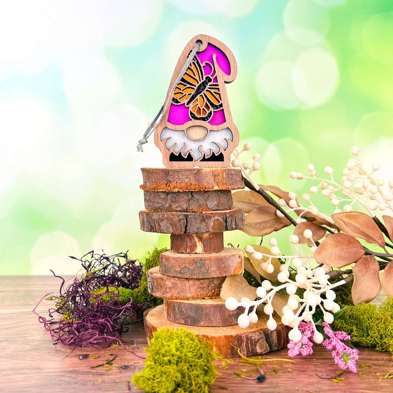 The Forged Flare® Monarch Butterfly Ornament, a 3.75" garden gnome with a pink hat from the Nature’s Gnomes™ collection, stands on stacked wood slices among dried flowers and greenery against a bright green background.