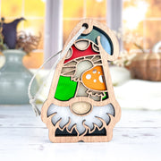 The Forged Flare® Toadstool Mushroom Ornament (3.75" H) from Nature's Gnomes™ features a bearded gnome with a red and green hat and mushroom accents, hanging by a string with blurred vases and an autumn window in the background.