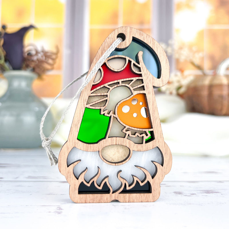 The Forged Flare® Toadstool Mushroom Ornament (3.75" H) from Nature's Gnomes™ features a bearded gnome with a red and green hat and mushroom accents, hanging by a string with blurred vases and an autumn window in the background.