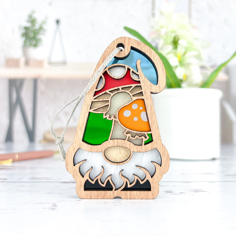 The Forged Flare® Toadstool Mushroom Ornament (3.75" H) from Nature's Gnomes™ features a garden gnome with a colorful, stained glass-style hat and mushroom design, displayed on a white tabletop with a blurred plant in the background.