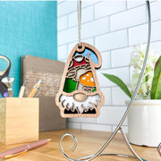 The Toadstool Mushroom Ornament | 3.75" (H) Garden Gnome Gift by Forged Flare® from Nature's Gnomes™ hangs on a silver stand on a desk with pens, a plant, and books against a tiled wall backdrop.