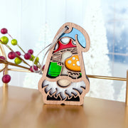 The Forged Flare® Toadstool Mushroom Ornament (3.75" H) from Nature's Gnomes™ features a wooden garden gnome with a white beard, blue hat, and colorful stained-glass mushrooms, hanging on a string. Festive berries and a frosted window blur in the background.