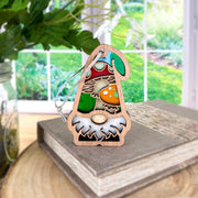 The Forged Flare® Toadstool Mushroom Ornament, a 3.75" Nature's Gnomes™ garden gnome gift, features a colorful gnome with a green hat and toadstool mushrooms sitting on a book, with a sunny garden in the background.