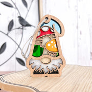 The Forged Flare® Toadstool Mushroom Ornament (3.75" H) from Nature's Gnomes™ features a wooden gnome with a white beard and red hat decorated with toadstools, hanging by a string against a leafy background on a light wood surface.