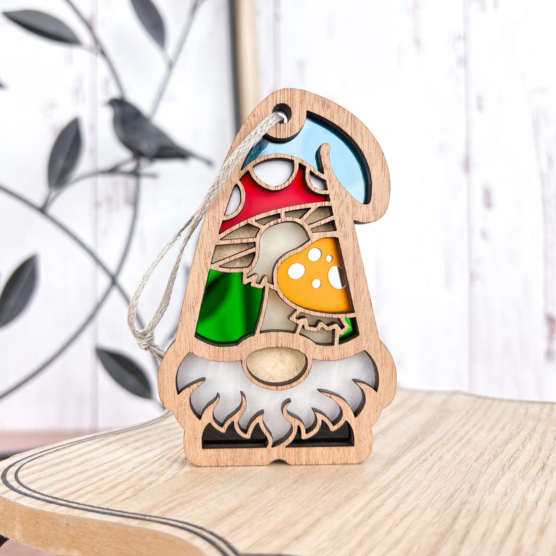 The Forged Flare® Toadstool Mushroom Ornament (3.75" H) from Nature's Gnomes™ features a wooden gnome with a white beard and red hat decorated with toadstools, hanging by a string against a leafy background on a light wood surface.