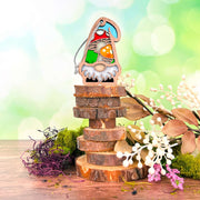 The Forged Flare® Toadstool Mushroom Ornament (3.75" H), from Nature's Gnomes™, features a colorful garden gnome standing on wooden slices with moss, faux berries, and leafy branches, set against a blurred green background.