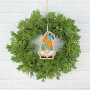 A green wreath on a white brick wall is adorned with the Forged Flare® California Poppy Ornament, a 3.75" Nature's Gnomes™ gnome holding an orange pumpkin and wearing an orange and blue hat.