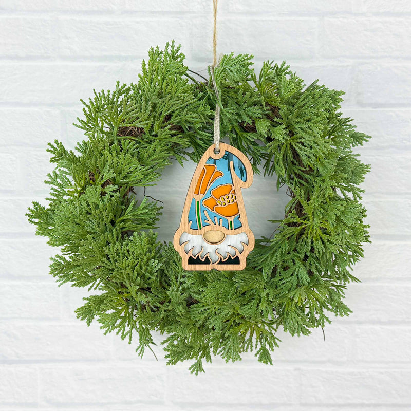 A green wreath on a white brick wall is adorned with the Forged Flare® California Poppy Ornament, a 3.75" Nature's Gnomes™ gnome holding an orange pumpkin and wearing an orange and blue hat.