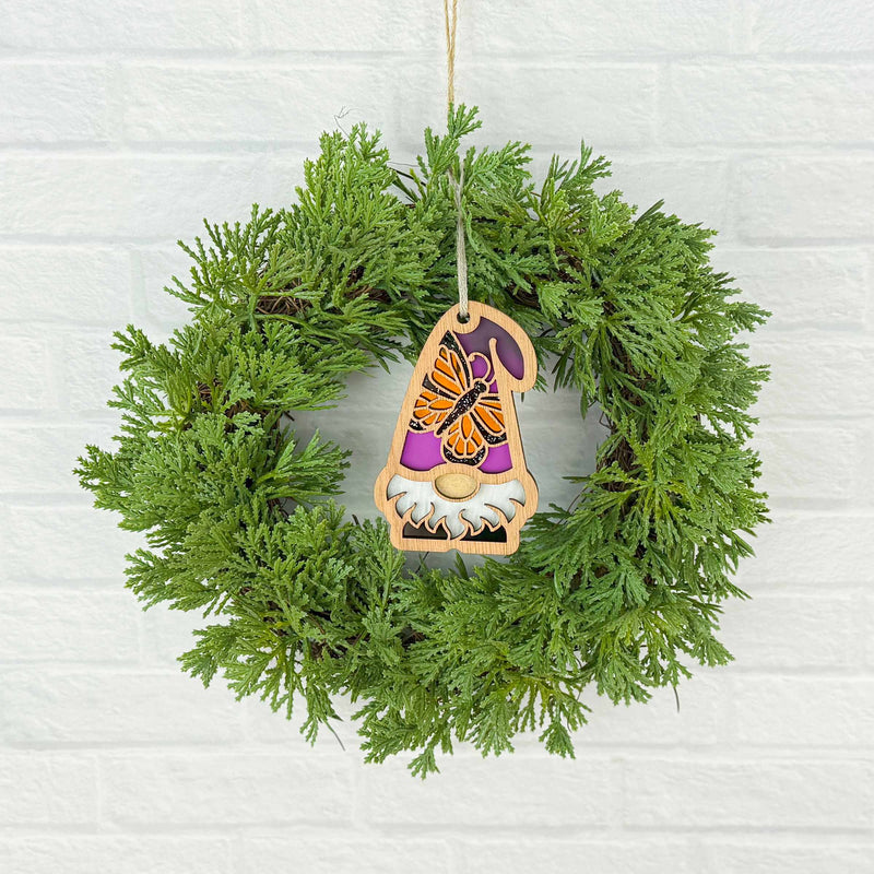 A green wreath hangs on a white brick wall, featuring the Forged Flare® Monarch Butterfly Ornament, a 3.75" purple-hatted gnome from the Nature's Gnomes™ collection.