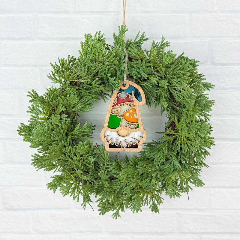 A green pine wreath hangs on a white brick wall, centered with Forged Flare®'s Toadstool Mushroom Ornament—a 3.75" garden gnome from the Nature's Gnomes™ collection holding a red toadstool mushroom.