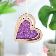 The Forged Flare® "I Love You" Purple Heart Ornament, 2.5" (H), features a sparkly glitter center and hangs on a white string, with blurred green leaves and pink flowers in the background.