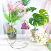 The Forged Flare® "I Love You" Purple Heart Ornament (2.5"H) sparkles with purple glitter on a metal stand, set against green plants and pink and white flowers on a white surface.