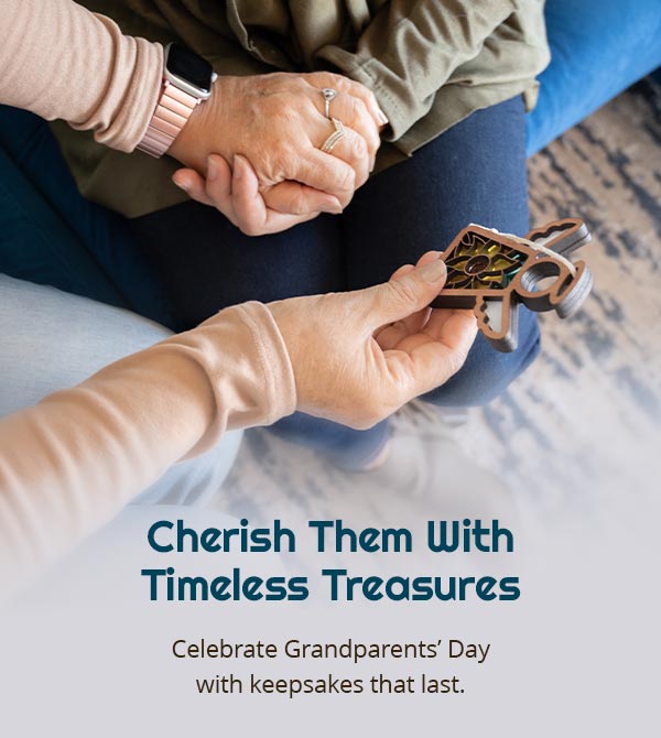 Cherish Them With Timeless Treasures. Celebrate Grandparents' Day with keepsakes that last. An intimate moment showing a grandchild gifting a Mother’s Angels® ornament to their grandparent. The image captures the sentiment and suggests thoughtful grandparents day gift ideas.