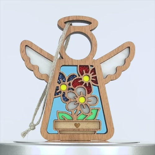 A wooden angel ornament with outstretched wings has a hollow circle for its head and a halo on top. Colorful, glittery flowers sit in a pot. A string is attached for hanging. The background is white, making it a perfect patriotic gift for any occasion.