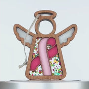 A wooden angel ornament with white wings and a pink ribbon on its front, surrounded by colorful confetti-like decoration. The angel has a braided linen string at the top for hanging, and a light string attached to the loop.