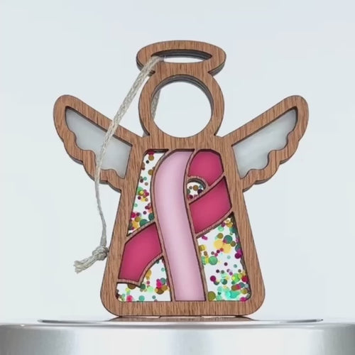 A wooden angel ornament with white wings and a pink ribbon on its front, surrounded by colorful confetti-like decoration. The angel has a braided linen string at the top for hanging, and a light string attached to the loop.