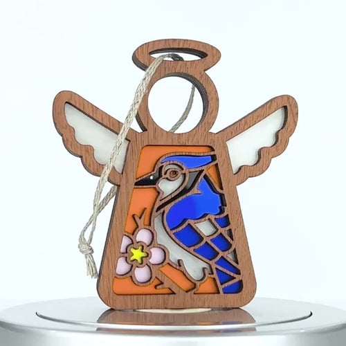 A wooden angel ornament showcasing a vibrant design of a blue jay and a pink flower. The angel has white wings and a circular halo with a string for hanging. The background features a mix of orange and blue hues, adding an artistic touch to any space.