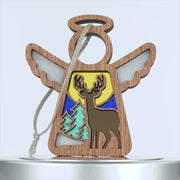 A wooden angel-shaped deer ornament features cutout details, including wings and a halo. The center depicts a deer against a colorful backdrop with green trees, a yellow sun, and a blue sky. A braided linen string is attached for hanging.