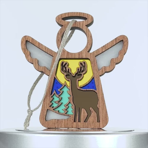 A wooden angel-shaped deer ornament features cutout details, including wings and a halo. The center depicts a deer against a colorful backdrop with green trees, a yellow sun, and a blue sky. A braided linen string is attached for hanging.
