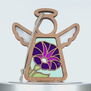 Wooden angel ornament with cutout wings and halo. Its center is adorned with a colorful, stained glass–style morning glory flower featuring purple and green petals. A loop of string is attached at the top for hanging. The background is white.