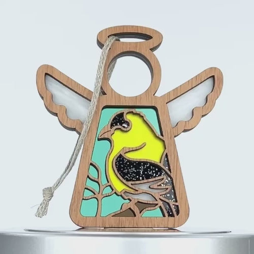 A wooden angel ornament with white wings is adorned with a colorful yellow and black goldfinch design on its body. The background is a light blue color, and a string is attached at the top for hanging.