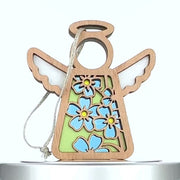 A wooden angel ornament with white wings features a green body adorned with blue and yellow forget-me-not flowers. The design includes a string loop for hanging. The angel's head and halo are carved out, making them hollow. The ornament is set against a white background.