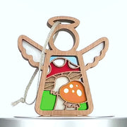 A wooden mushroom ornament in the shape of an angel. The angel has a halo and wings, with a string for hanging. The red and orange mushrooms with white spots are set against a backdrop of blue and green.