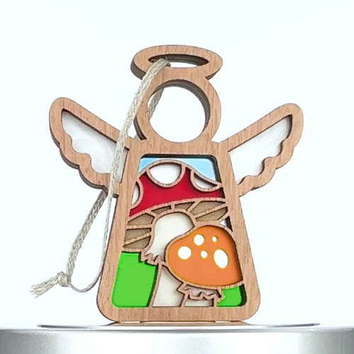A wooden mushroom ornament in the shape of an angel. The angel has a halo and wings, with a string for hanging. The red and orange mushrooms with white spots are set against a backdrop of blue and green.