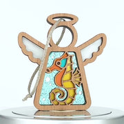 A wooden angel seahorse ornament with transparent wings and a halo. The body features a stained glass–style design depicting a colorful seahorse with orange, teal, and yellow hues. The ornament hangs from a braided linen string.