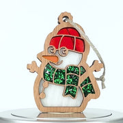 A wooden snowman ornament with cut-out detailing. The snowman features an orange carrot nose, a red hat, and a green glittery scarf. A braided linen string is attached at the top for hanging.