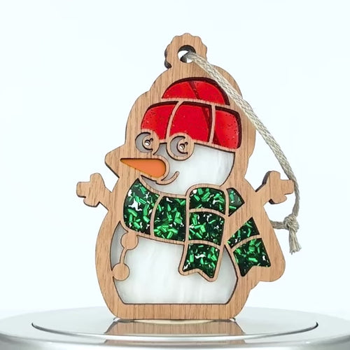 A wooden snowman ornament with cut-out detailing. The snowman features an orange carrot nose, a red hat, and a green glittery scarf. A braided linen string is attached at the top for hanging.