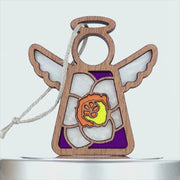 A wooden angel ornament with its wings outstretched, featuring a stained glass–style design. The body has purple and white details, with a central yellow flower reminiscent of a jonquil. A braided linen string is attached at the top for hanging. The ornament is set against a white background.