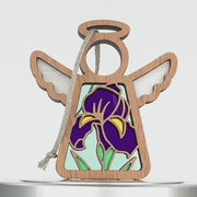 A decorative wooden angel with open wings and a round halo features a colorful stained glass–style design of purple irises and green leaves, making it a charming iris ornament. A braided linen string is attached at the top for hanging, set against a plain white background.