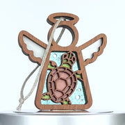 A wooden angel-shaped ornament with carved wings and a halo, featuring a sea turtle in the center. The turtle has a brown shell with green accents and is surrounded by a blue background with bubble-like patterns. A braided linen string is attached to the ornament.